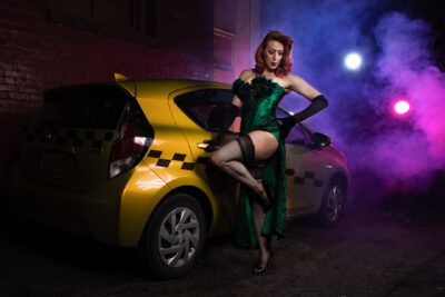 A person dressed in a green corset, black stockings, and long black gloves poses next to a yellow car with a checkered pattern on the side, reminiscent of a taxi. The scene includes fog and colorful lighting with blue and purple hues, creating a dramatic atmosphere.