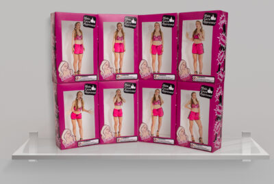 A display shelf holds six identical dolls in pink packaging. Each box shows a blonde-haired doll wearing a pink outfit and heels. The packaging features the text "Male Gaze Approved" and a pink silhouette of a woman's profile on the side. The dolls are arranged in two rows.